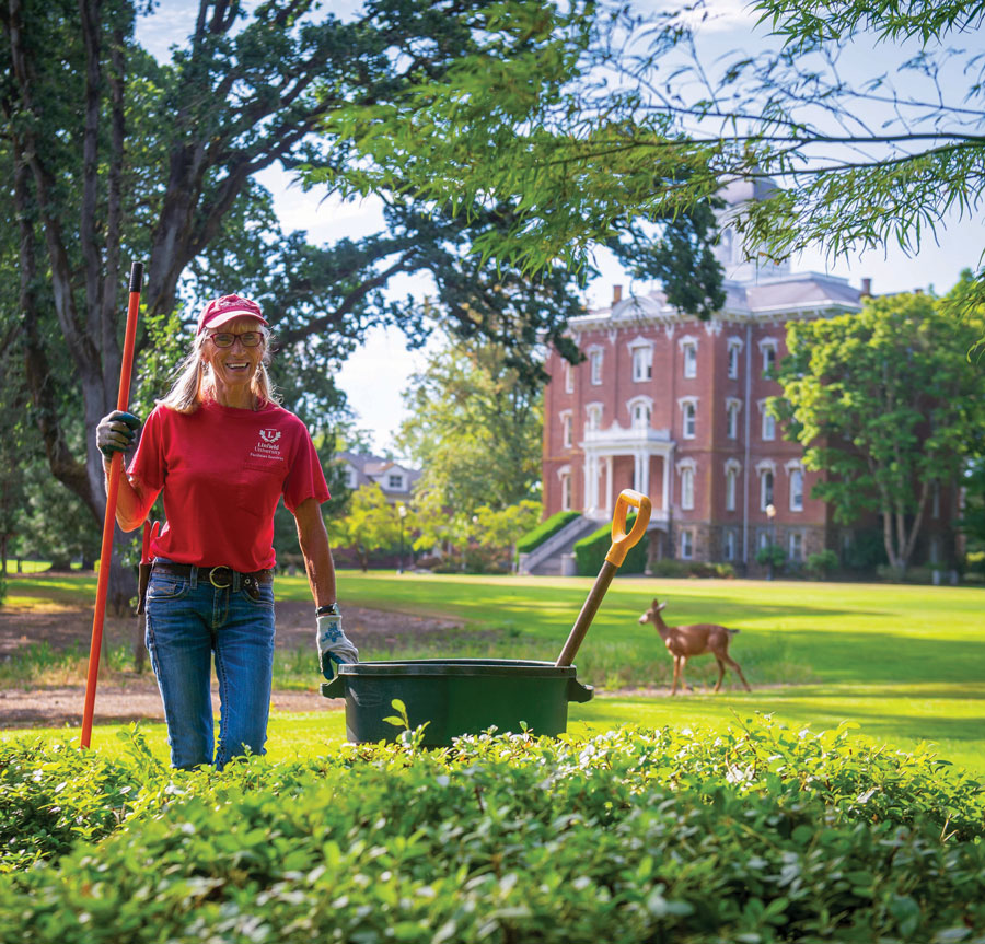 Carol Gallagher working in the Oak Grove with a deer and Pioneer Hall behind her
