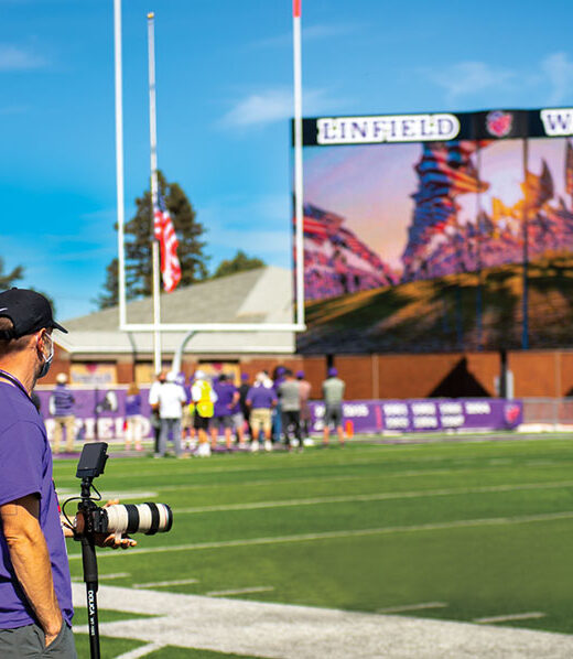 Mike Lempner, video producer, on the sidelines of the football field with the video screen in the background