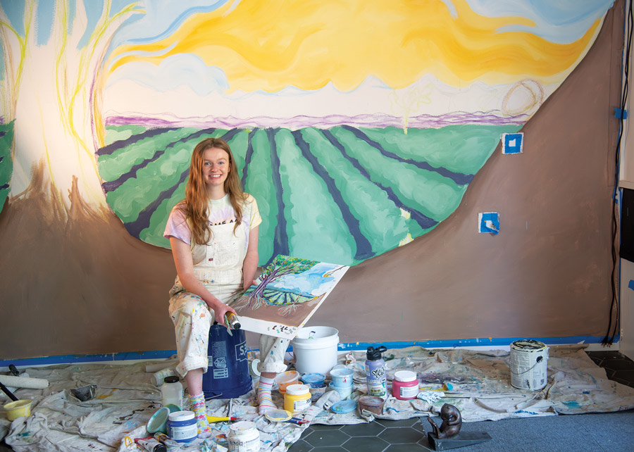 Linfield student Alliso Hmnura '26 sitting on a bucket in front of her mural at Acorn to Oak.