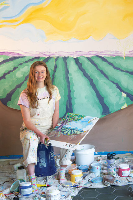Linfield student Alliso Hmnura '26 sitting on a bucket in front of her mural at Acorn to Oak.