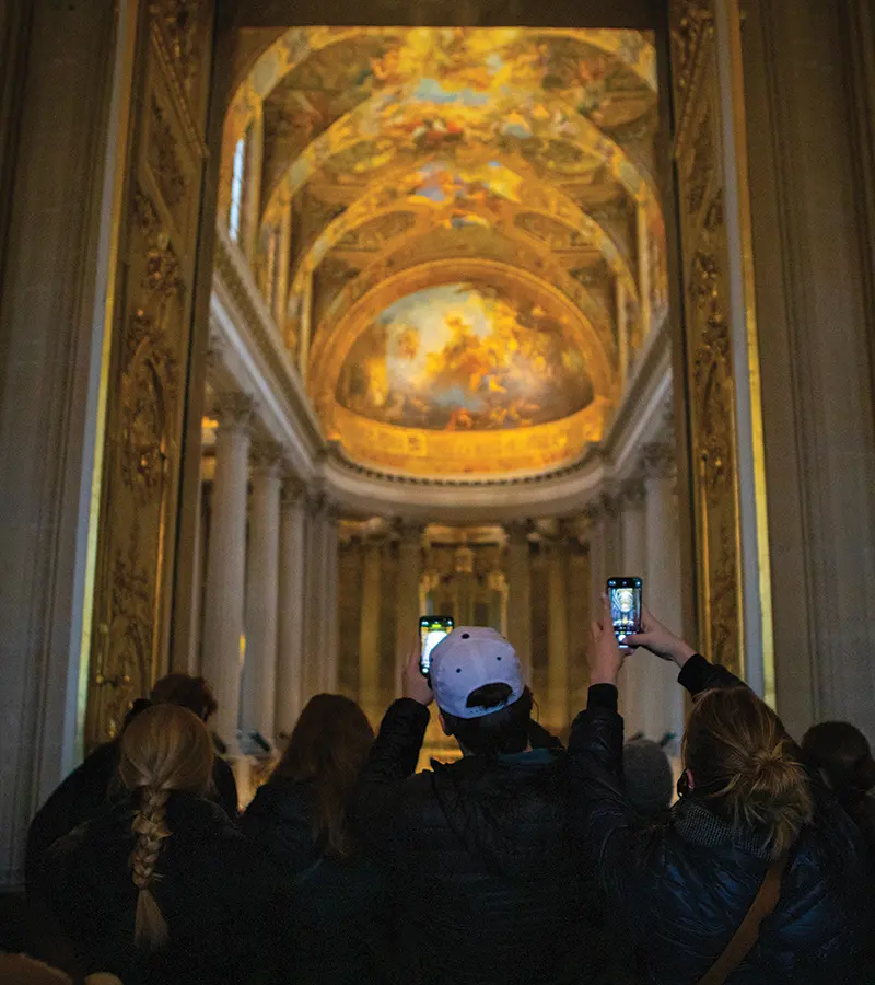 Students visit the Palace of Versailles and take photos of the ceiling with their phones.