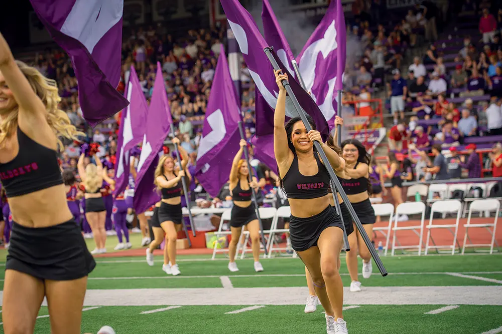 The cheer team runs onto the football field holding flags.