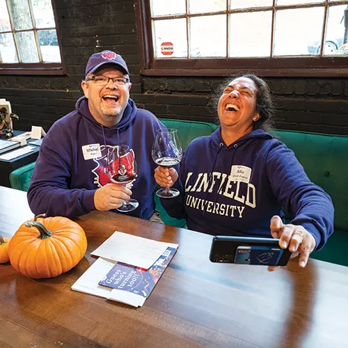 two people sitting at a table, laughing while holding wine glasses and taking a selfie.