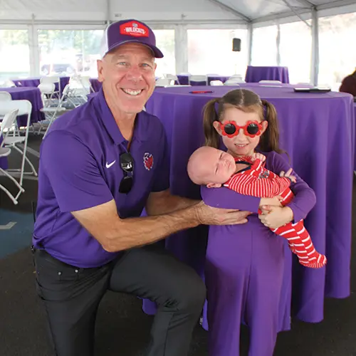 Athletic Director Scott Brosius posing for a photo with a child who is holding a baby.