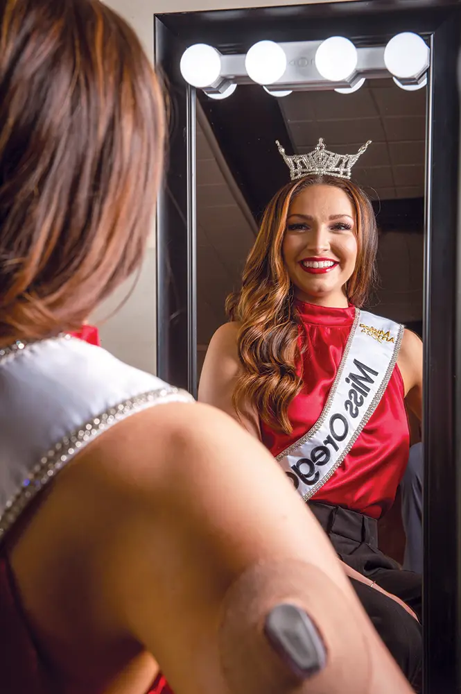 Abigail looking in a mirror while wearing her Miss Oregon sash and crown.